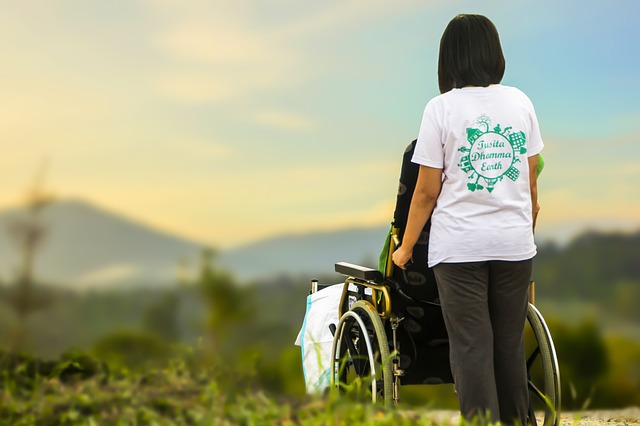 Osoba niepełnosprawna na wózku inwalidzkim, spacerująca na łące i prowadzona przez opiekuna