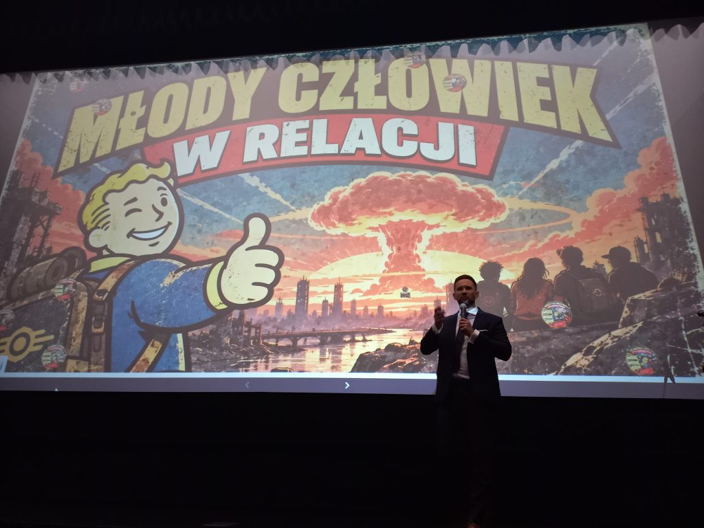 Mężczyzna w ciemnym garniturze stoi na scenie, przemawiając do publiczności przez mikrofon. Za nim na dużym ekranie wyświetlana jest grafika z napisem „Młody człowiek w relacji”, przedstawiająca postapokaliptyczny krajobraz z wybuchem atomowym oraz uśmiechniętą postać z gry Fallout, pokazującą uniesiony kciuk.