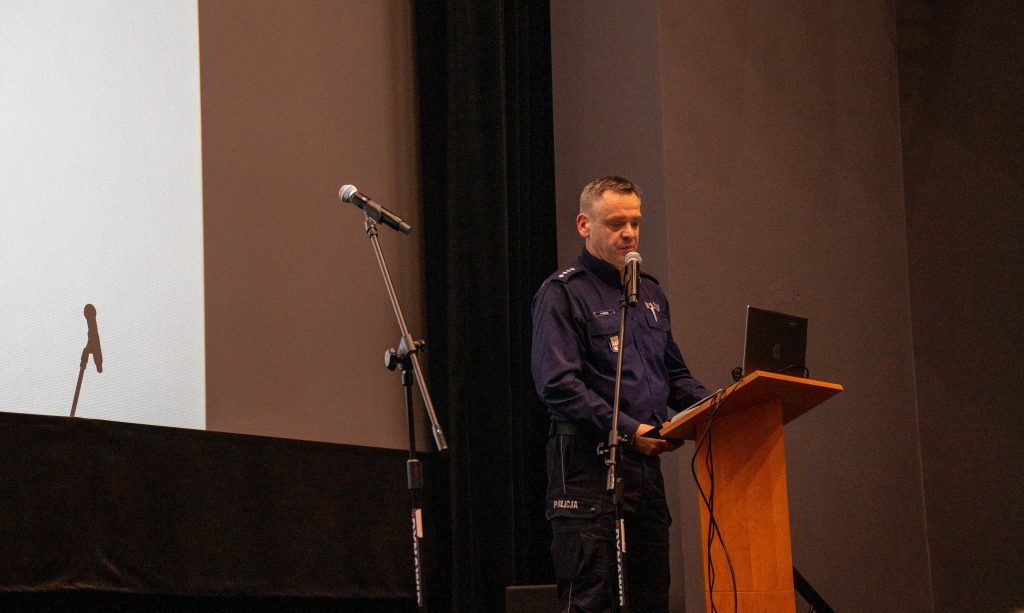 Policjant w granatowym mundurze przemawia do publiczności na scenie, stojąc za drewnianą mównicą z laptopem. Mówi do mikrofonu umieszczonego na statywie. Obok znajduje się drugi mikrofon, a w tle widoczny jest fragment ekranu projekcyjnego.