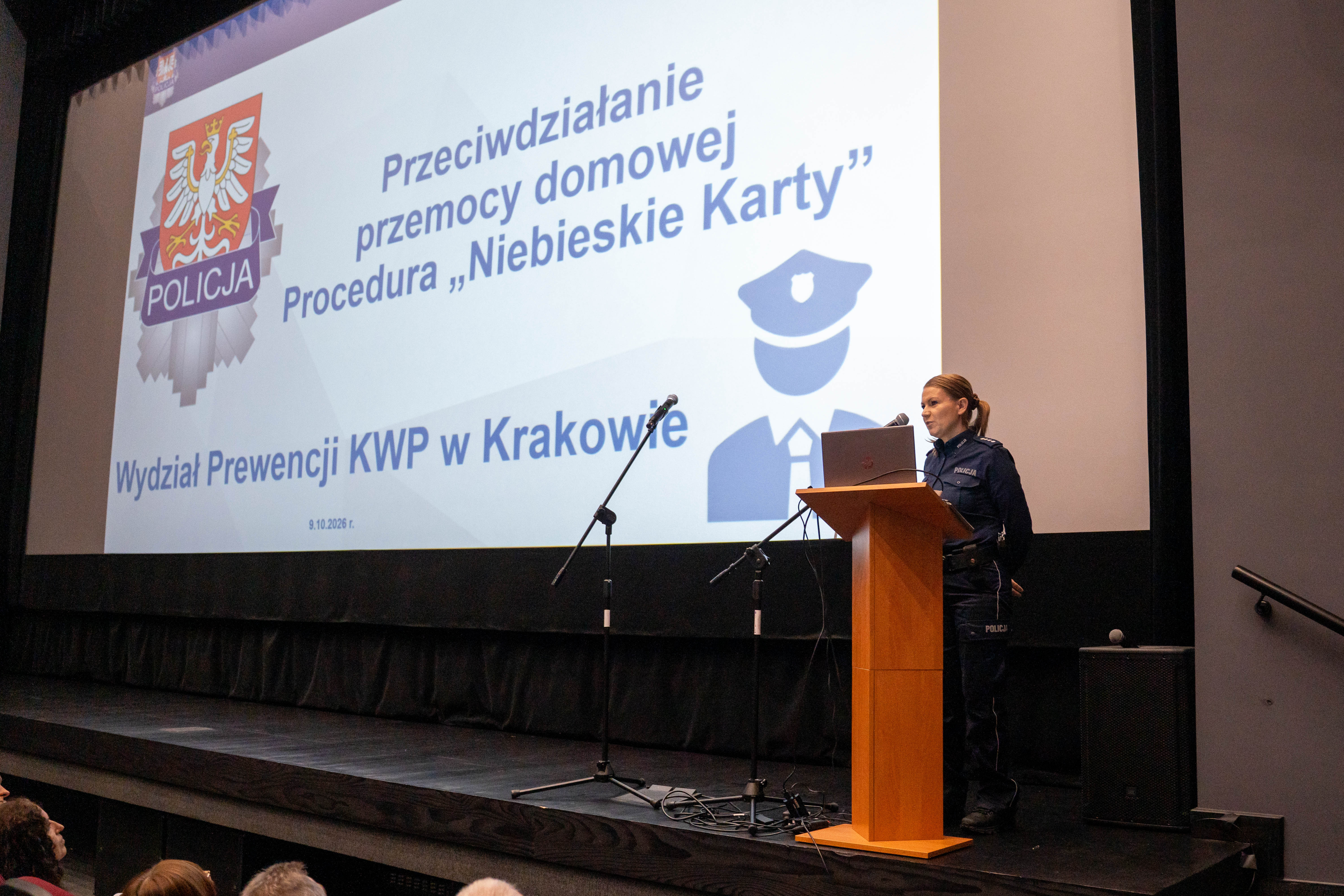 Policjantka w mundurze prowadzi prezentację na scenie, stojąc przy drewnianej mównicy z laptopem. Za nią wyświetlany jest slajd Komendy Wojewódzkiej Policji w Krakowie zatytułowany „Przeciwdziałanie przemocy domowej. Procedura Niebieskie Karty”. Na pierwszym planie widoczni są uczestnicy spotkania.