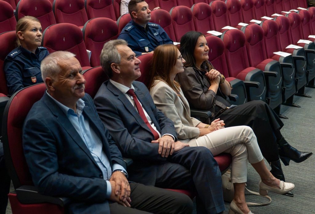Uczestnicy spotkania siedzą w bordowych fotelach sali kinowej, obserwując wydarzenie na scenie. W pierwszym rzędzie znajdują się dwaj mężczyźni w garniturach oraz dwie kobiety w eleganckich strojach. Za nimi siedzi dwoje funkcjonariuszy policji w granatowych mundurach.