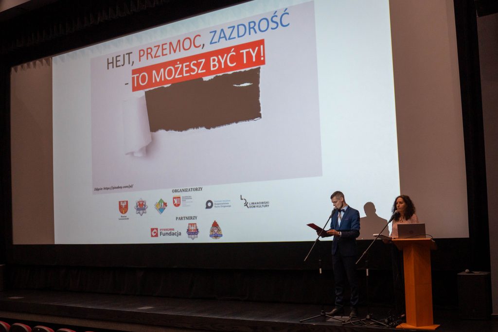 Na scenie sali kinowej mężczyzna w granatowym garniturze oraz kobieta stojąca przy drewnianej mównicy prowadzą spotkanie, przemawiając do mikrofonów. Za nimi na dużym ekranie wyświetlany jest plakat wydarzenia z hasłem „Hejt, przemoc, zazdrość – to możesz być Ty!” oraz logotypami organizatorów i partnerów.