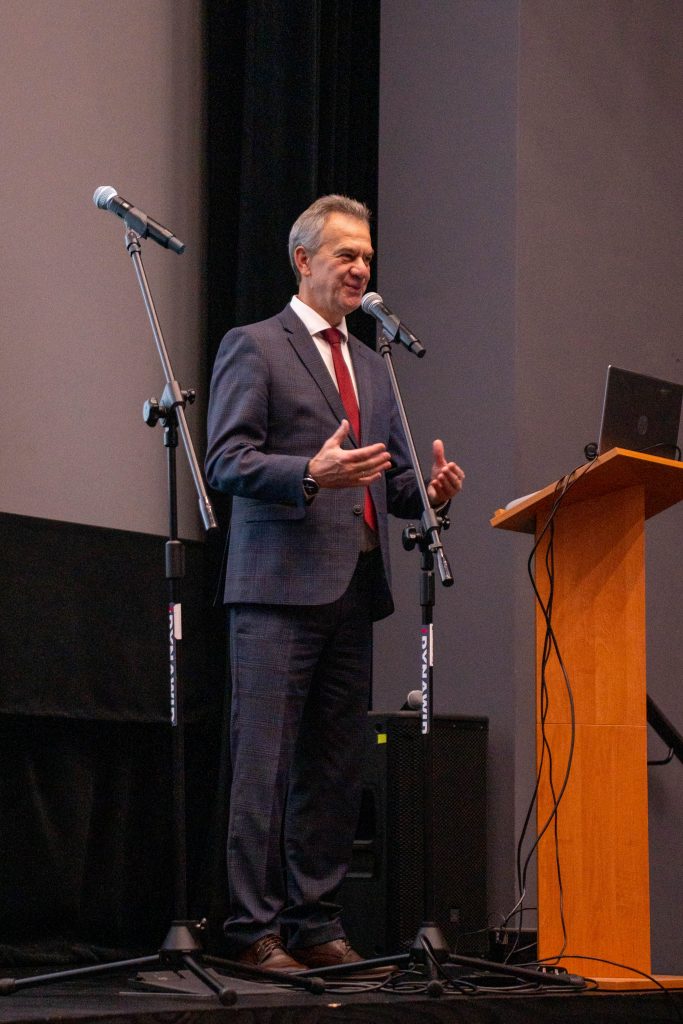 mężczyzna w granatowym garniturze, białej koszuli i czerwonym krawacie przemawia do publiczności na scenie. Stoi przed mikrofonem na statywie, gestykulując obiema rękami. Obok znajduje się drewniana mównica z laptopem.